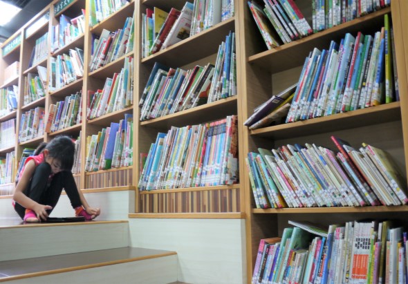 一名穿著黑色上衣和粉紅色鞋子的學生坐在圖書館地板上，身後是裝滿各色書籍的木製書架。書架整齊排列著數百本書籍，展示了豐富的圖書館藏書。