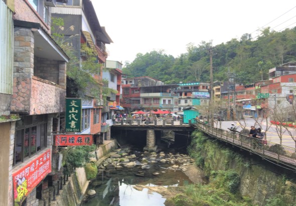 山區小鎮街景，兩岸建築密集，紅色和彩色房屋沿著溪流排列。橋樑跨越水道，綠色植被覆蓋周圍山坡，街道上有商店招牌和行人，呈現典型的中國南方農村聚落風貌。