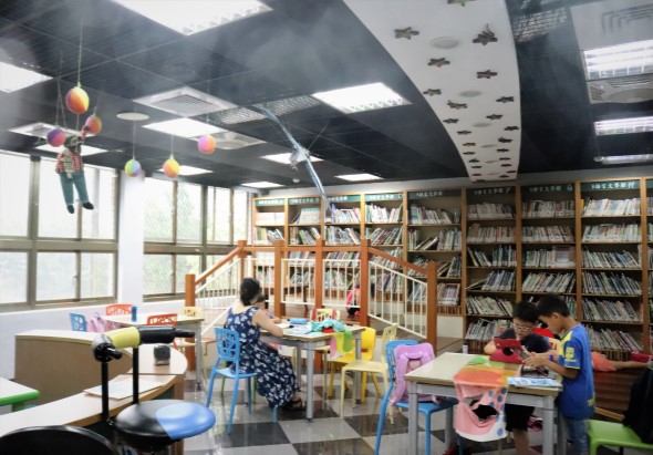 現代圖書館兒童閱讀區，寬敞明亮的空間配備木製書架、彩色閱讀桌椅和遊戲區。數名兒童正在進行閱讀和學習活動，天花板懸掛著五彩繽紛的裝飾品，營造溫馨友善的學習環境。