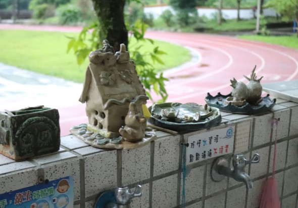 公園水池邊的石造欄杆上，擺放著多個陶製裝飾品，包括古式房屋、動物造型和植物盆景。背景可見綠色草坪、樹木及紅色跑道。