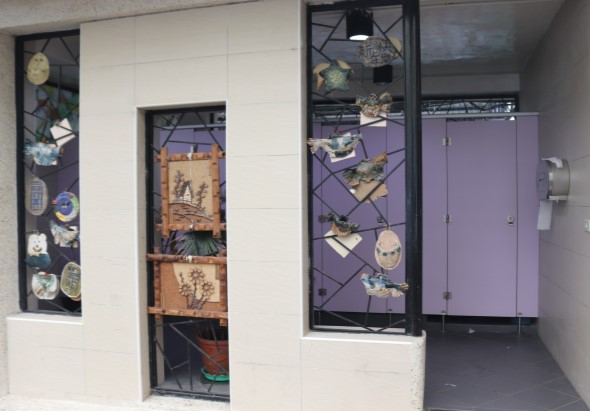現代藝術展示空間入口，左側白牆上方有豎形黑色展示櫃展示陶瓷盤碟，中央玻璃門展示木製掛飾和陶器，右側高展櫃陳列各式陶藝作品。紫色門框與白色牆面形成簡潔展覽環境。