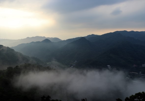 晨霧籠罩的山谷景觀。層疊的青色山巒在晨光中逐漸淡去，山谷間瀰漫著白色霧氣，隱約可見河流和零星建築。天空呈現淡藍色，陽光從左側透出，營造出寧靜而夢幻的山林氛圍。