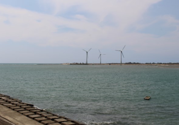 海邊風景，前景是防波堤，中景是平靜的青綠色海水，遠景是陸地上矗立的三座白色風力發電機，天空為陰云密布。一艘小船停靠在海面上。