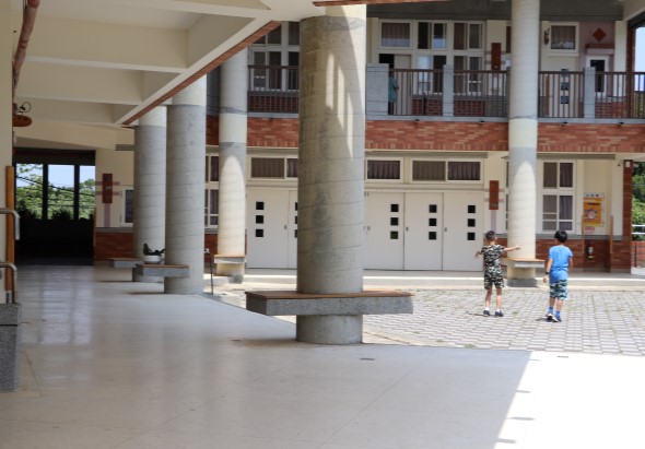 寬敞的學校或公共建築室內廣場，地面光滑平坦。多根灰色圓形支柱支撐上層結構，紅磚牆面可見。兩名兒童穿著運動服在場地中央活動。背景有白色大門、樓梯和上層走廊。陽光透過左側窗戶灑入。