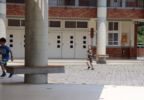 學校操場上，兩名穿著運動服的小學生在奔跑玩耍。背景是紅磚白色校舍建築和灰色支撐柱。地面鋪設灰色方形磁磚，光線充足的戶外場景。