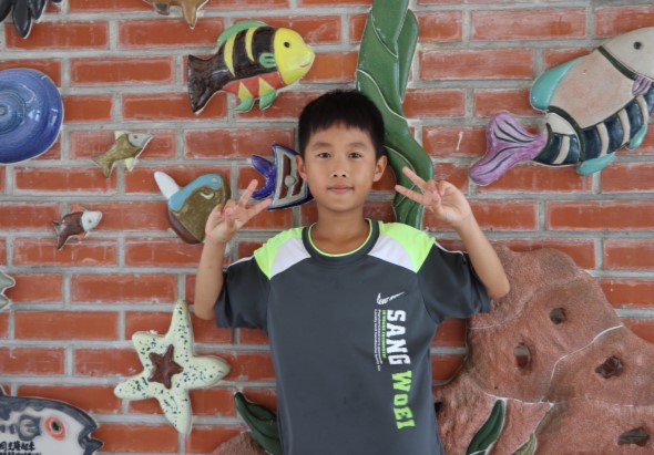 一名穿著Nike運動服的男孩在海洋生物主題的紅磚牆壁前擺姿勢。背景裝飾有彩色海魚、海星和海草剪貼畫，男孩雙手舉起做出歡樂手勢，臉帶微笑。