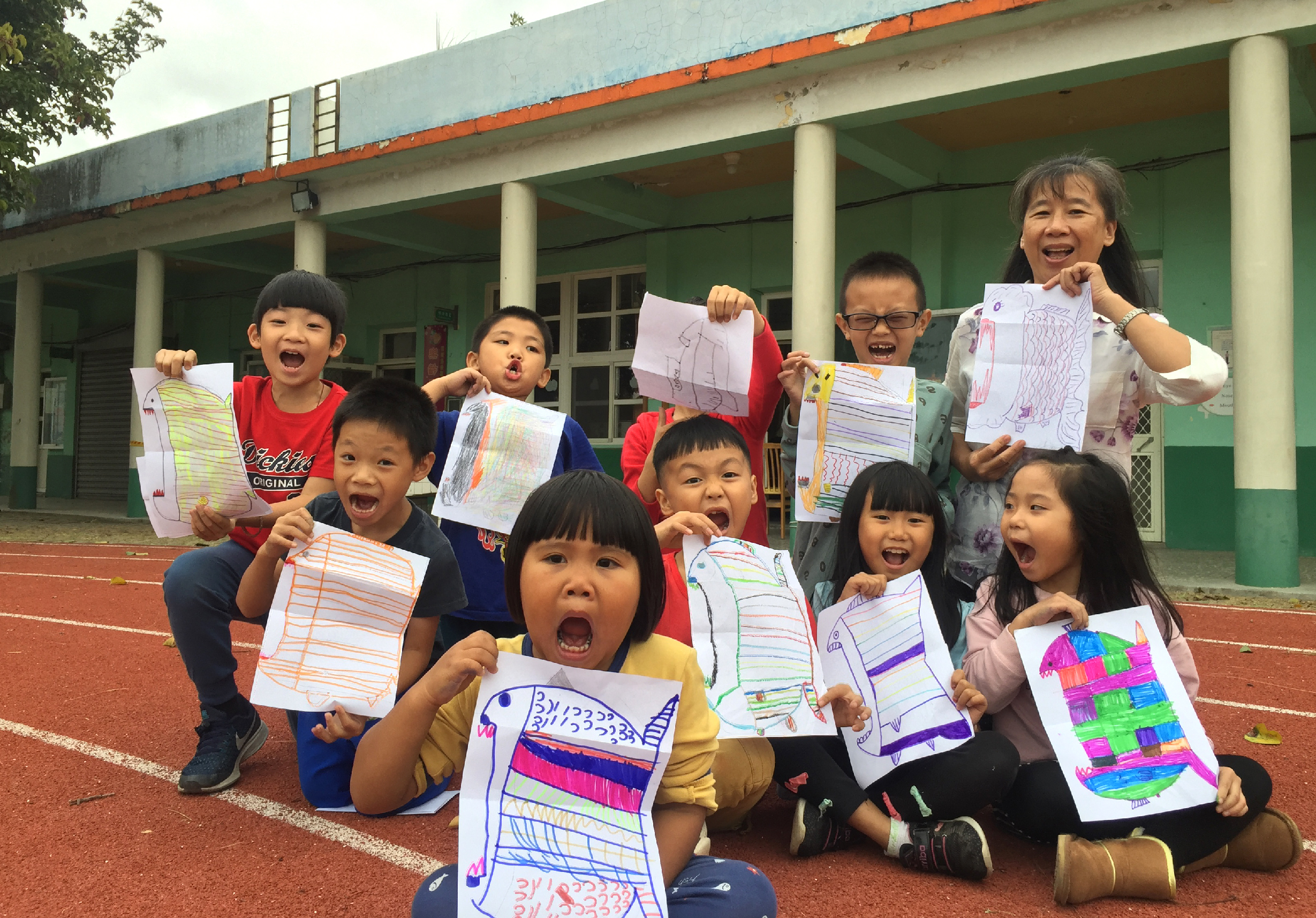 一群小學生在校園紅磚跑道上開心地展示自己的美術作品。孩子們穿著不同顏色的衣服，舉著畫有線條、色彩繽紛的紙張，在綠色校舍前燦爛笑容，表現出完成作品的喜悅。