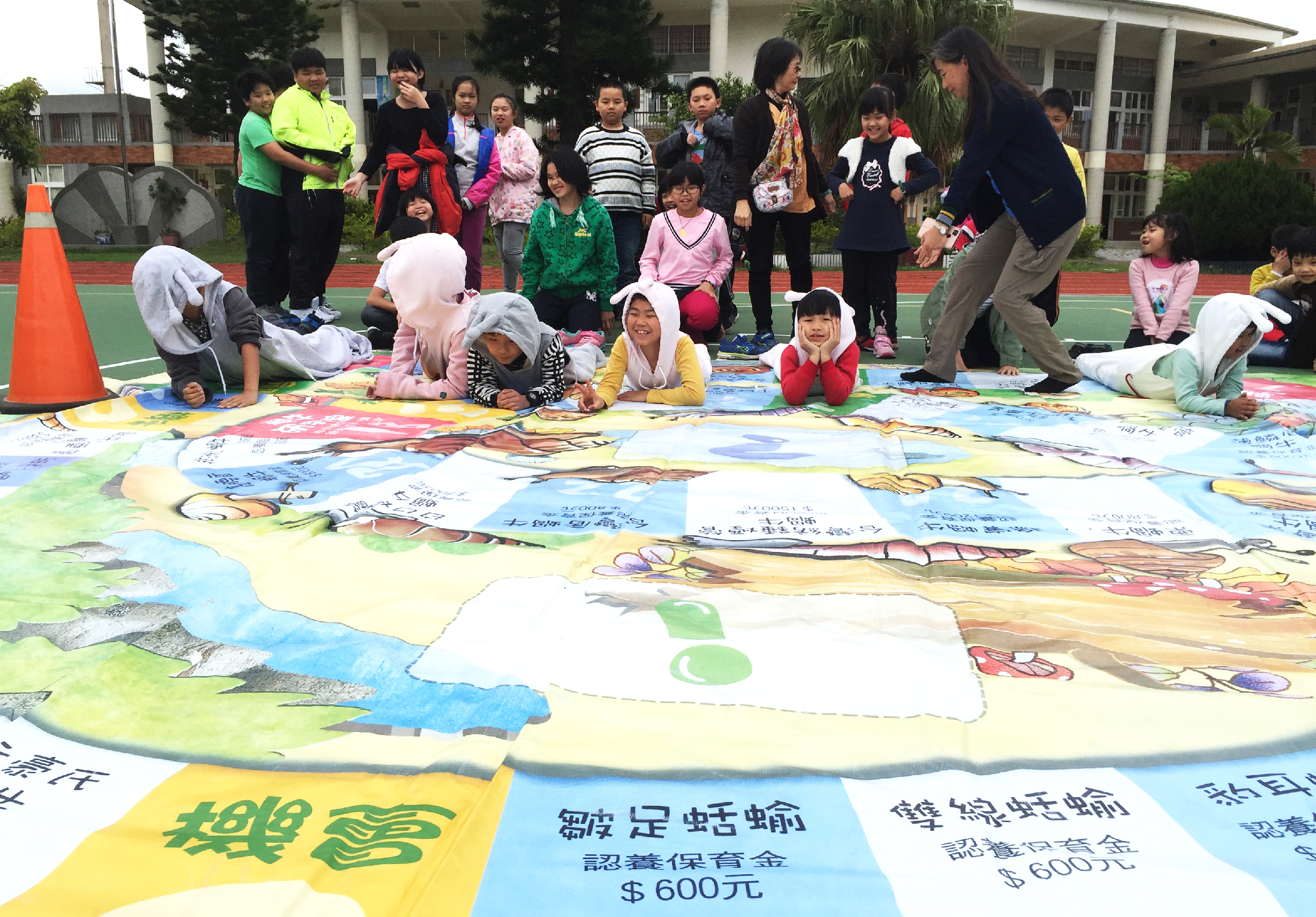 學校操場上舉辦兒童繪畫展覽活動。大量五彩繽紛的兒童畫作鋪滿地面，展示山水風景、人物等主題。數十名小學生圍坐或站立觀賞，教師在旁指導。黃色和藍色標語牌標註作品資訊和募款金額。背景為校舍和綠化環境。