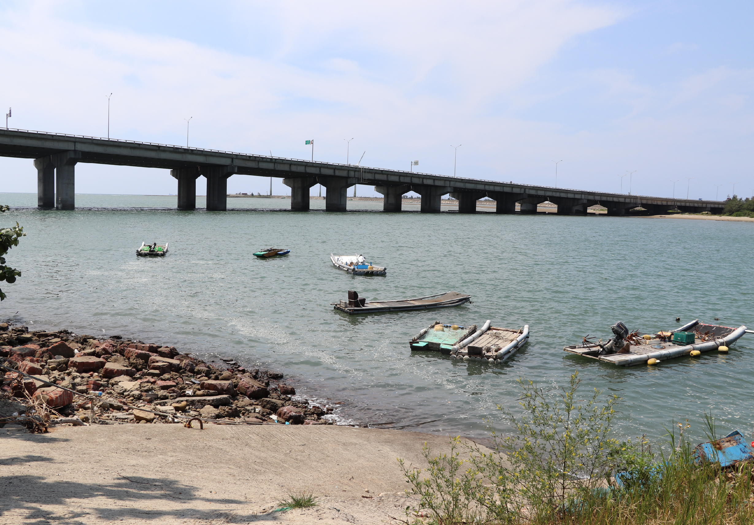 海邊沙灘靠岸，多艘小漁船停泊在淺藍色海水中。遠方跨越海灣的長橋由多根支柱撐起，橋面筆直延伸。岸邊散落紅褐色石塊，灘上生長稀疏草木。晴朗天空映照波光粼粼的海面，呈現寧靜漁村景象。