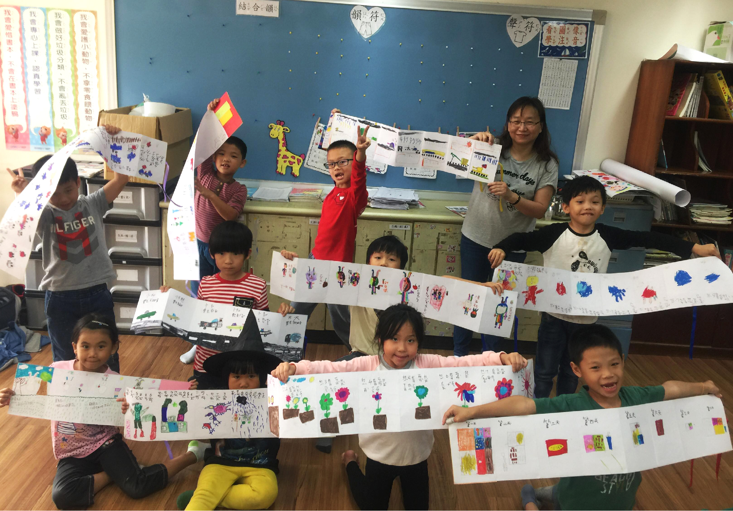 小學教室內，約十位學生舉著自己繪製的長條形畫作，展示色彩繽紛的花卉、植物和創意圖案。學生們穿著各色衣服，開心地笑著。教室牆面布置著藍色底板、心形卡片和各式學生作品，營造溫暖的學習環境。