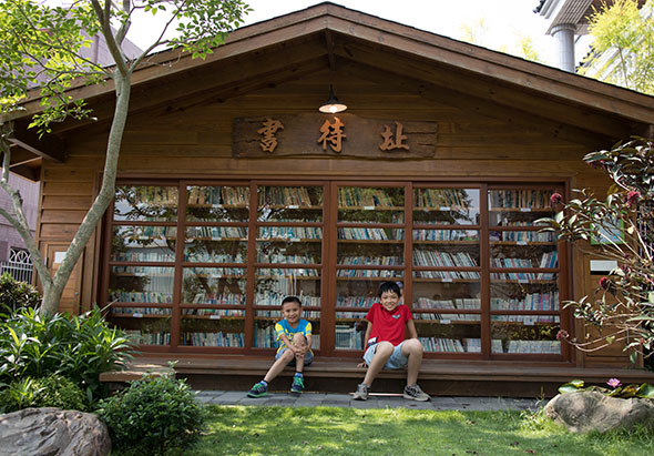 木製書屋外，兩個男孩坐在草地上閱讀。建築物正面有六扇玻璃書櫃窗，擺滿書籍。屋頂懸掛木製匾額，周圍環繞綠樹和植物，營造寧靜的閱讀環境。