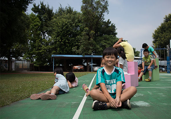 在戶外遊樂場，一名穿著綠色運動衣的小女孩坐在綠色地面上微笑，身後有其他兒童在玩耍。場景中有粉紅色遊樂設施、樹木和藍色棚子，是一個充滿歡樂的兒童活動現場。