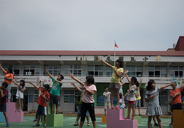 小學校園內，眾多學童站在彩色方形台子上做伸展動作，背景是紅頂校舍。孩子們穿著運動服，在戶外進行集體體操或健身活動，展現活力與朝氣。