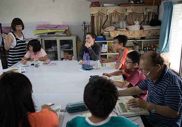 教室內進行閱讀教學活動，約八位成人和兒童圍坐在白色長桌前，每人持有課本。左側站立的女性講師正進行教學，黑板、書架和教學用具分別布置在教室各處。