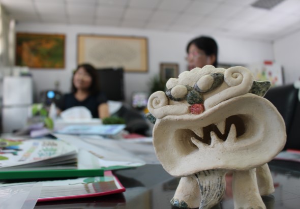 辦公室內，前景中有一尊白色陶瓷雕像，造型為張嘴的怪獸或神獸形狀。背景模糊可見兩位員工在桌前工作，桌上散放著文件、筆記本和辦公用品，牆上掛著畫作和地圖。