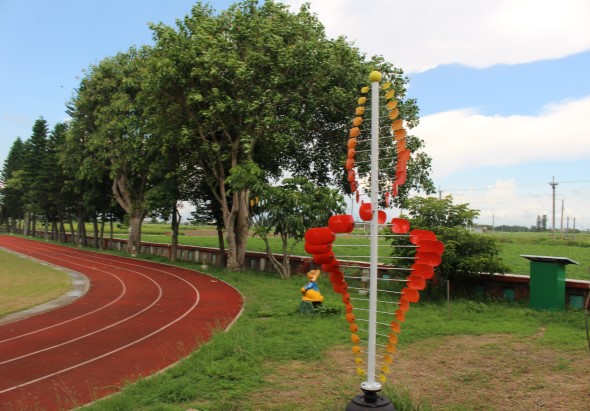 公園內紅色跑道旁，一根白色柱子上裝飾著紅色和橙色的花卉布置，形成愛心圖案。背景可見大樹、綠色草坪和遠處的山脈。一名兒童在柱子附近活動。
