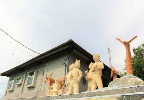 一棟灰白色建築物前設有多座金色雕像，包括兩個展開雙臂的人物雕像和兩個橙紅色的鳥類雕像。背景可見電塔和綠色植被。