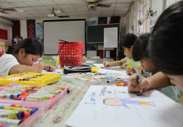 教室內，四名小學生圍坐在長桌旁進行美術課程。他們正在用彩色筆和標記筆繪畫，桌上散放著多色彩筆、畫紙和美術用具。教室後方有白板、黑板和儲物架。
