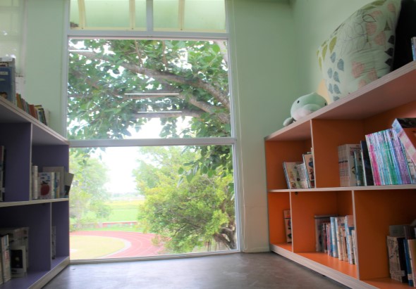 明亮的圖書室內景，左側為深藍色書架，右側為橙色書架上放滿各類書籍。大型落地窗採光充足，窗外可見綠樹和運動場。室內擺有球類裝飾品，營造溫馨的閱讀環境。