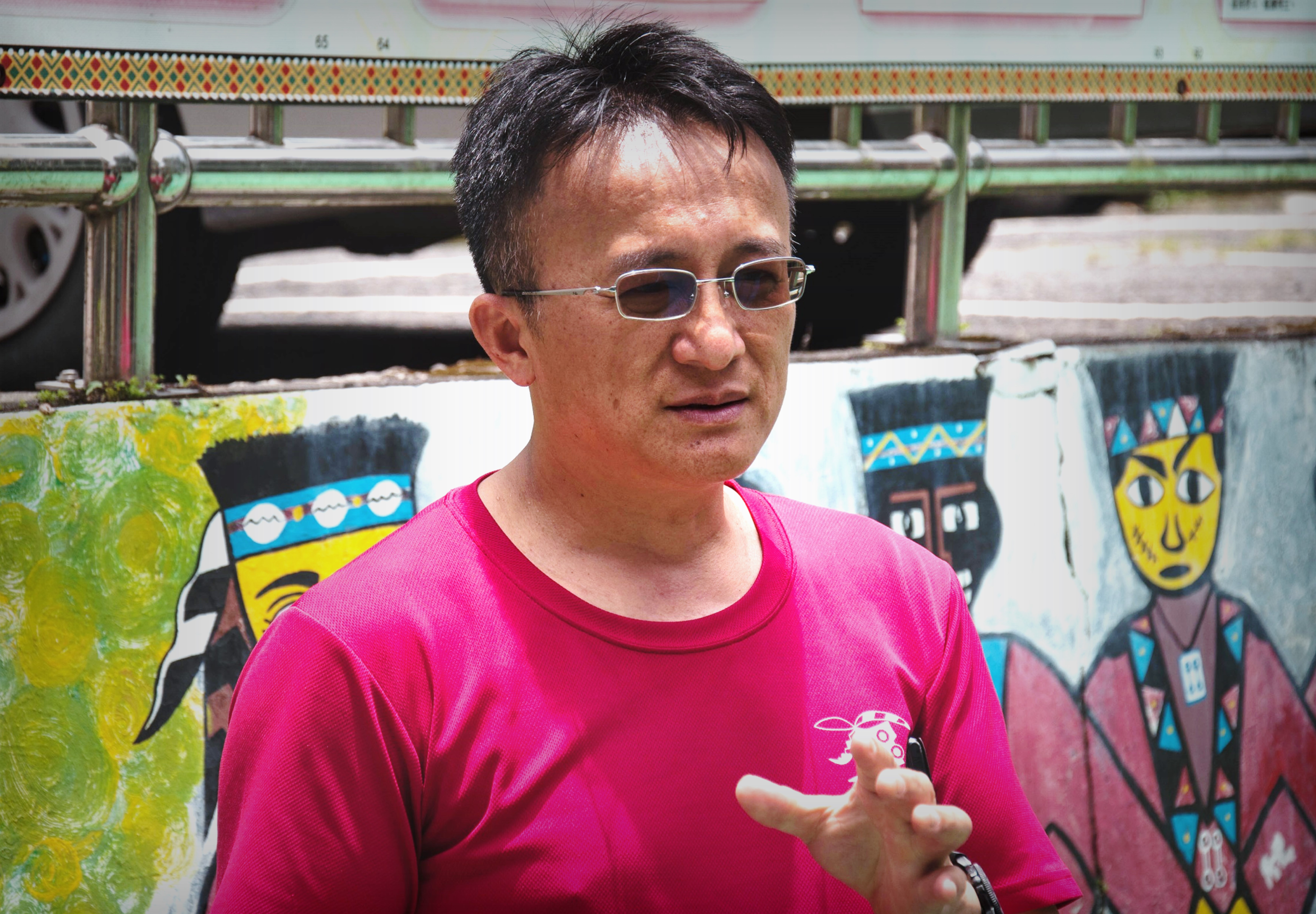 一位穿著桃紅色T恤、戴著眼鏡的男性在戶外進行講解或演講。背景是彩色塗鴉牆面和工業建築結構。他做出手勢動作，表情專注認真，似乎正在進行教學或介紹活動。