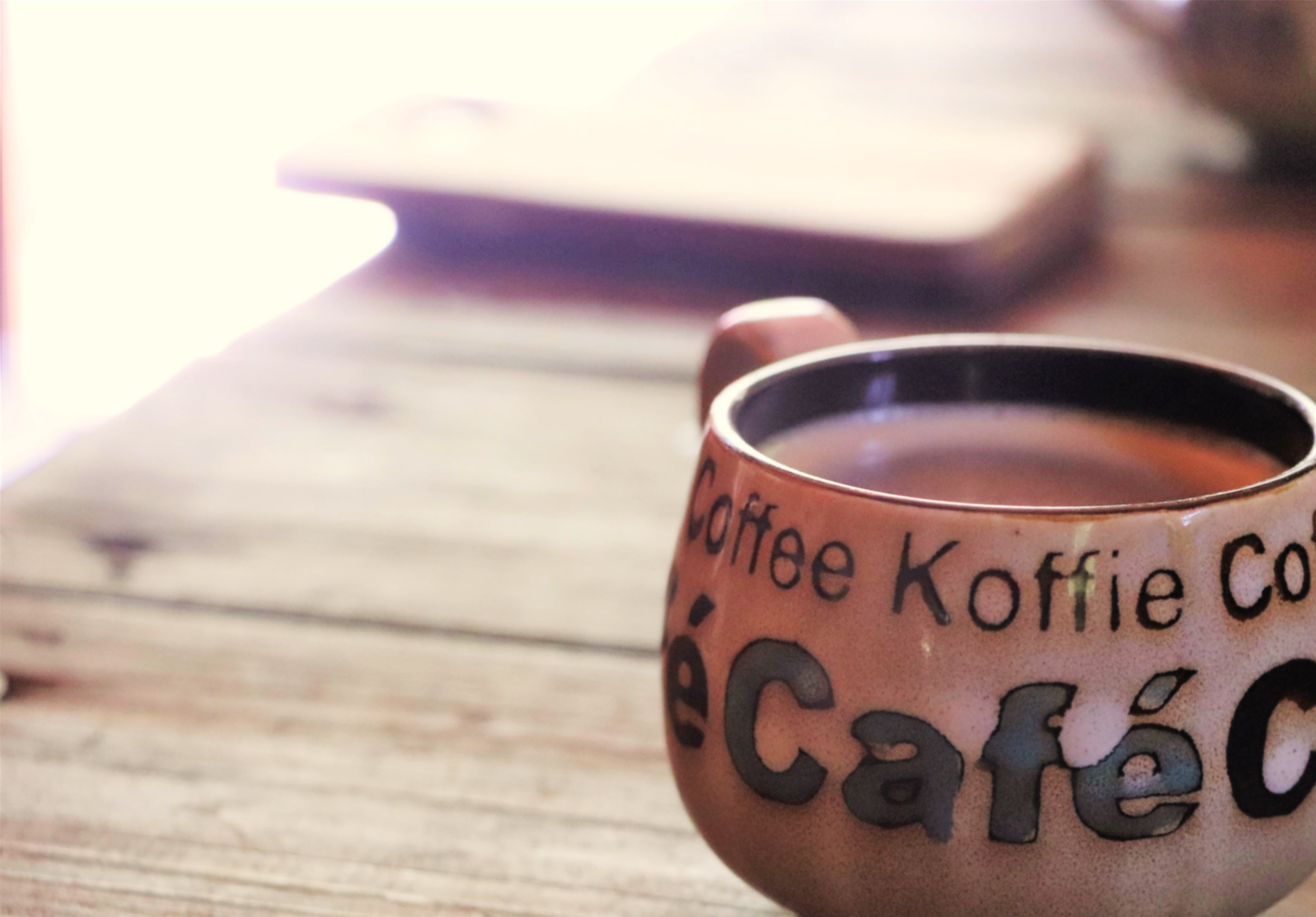 木質桌面上放著一個棕色馬克杯，杯身印有「Coffee Koffie Co」和「Café」字樣，杯中盛著熱咖啡。背景模糊可見筆記本和其他物品，營造舒適的咖啡時光氛圍。