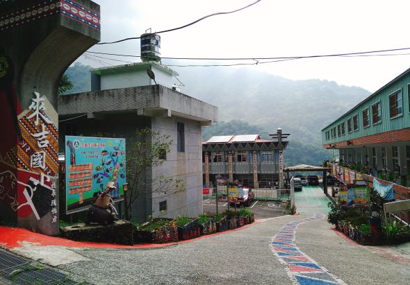 山區村落廣場，彩虹色地面彩繪，周圍有紅色壁畫建築、綠色建築物，遠處群山籠罩霧氣。廣場中央有裝飾花卉和指示牌，呈現鄉村藝術創意街區景象。