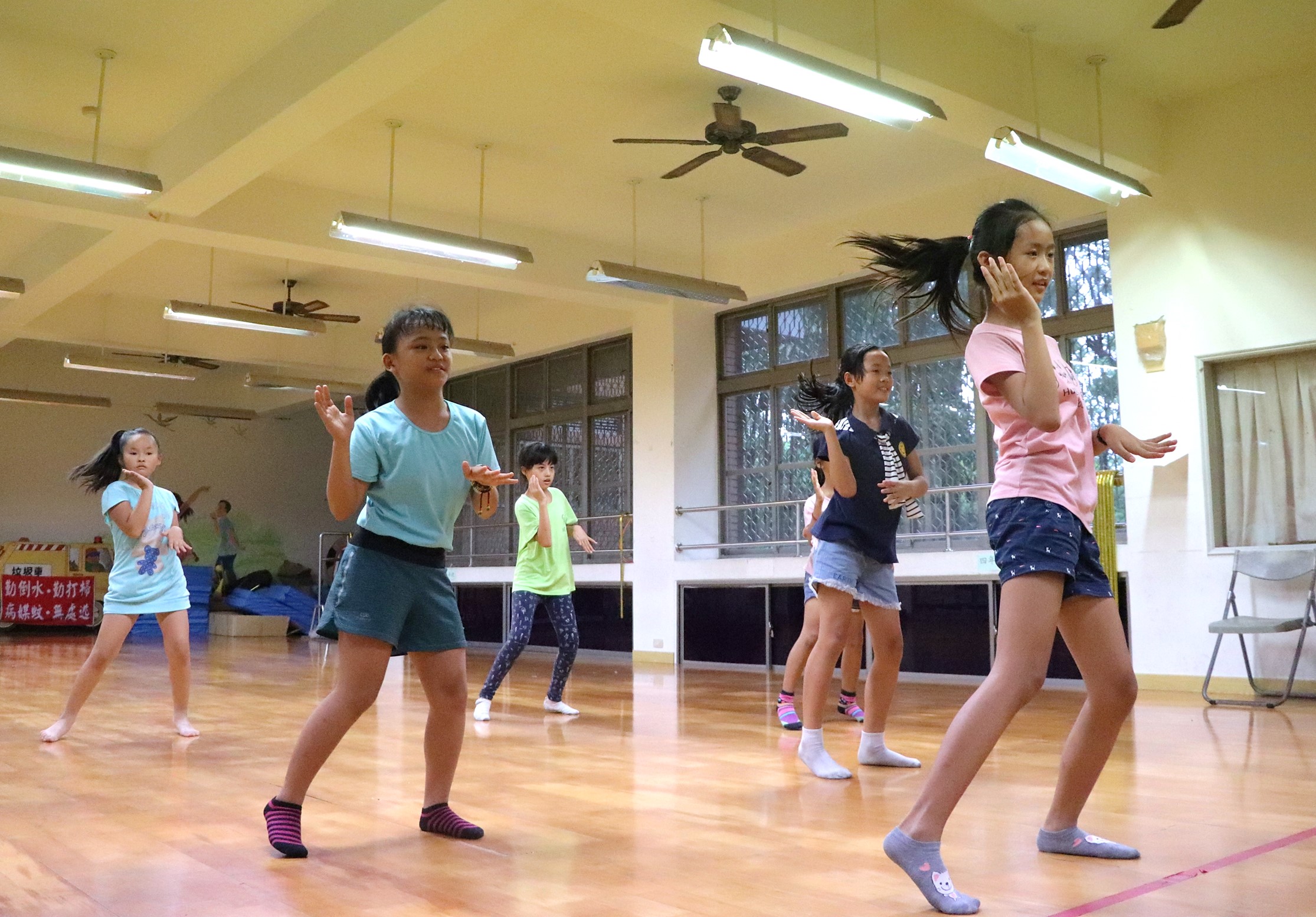 一群小學女童在體育館內進行跳繩運動課程。寬敞明亮的室內場地配有木質地板、天花板吊扇與日光燈。多位女童穿著運動服裝，在教練帶領下認真練習跳繩動作，展現活力與專注的運動精神。