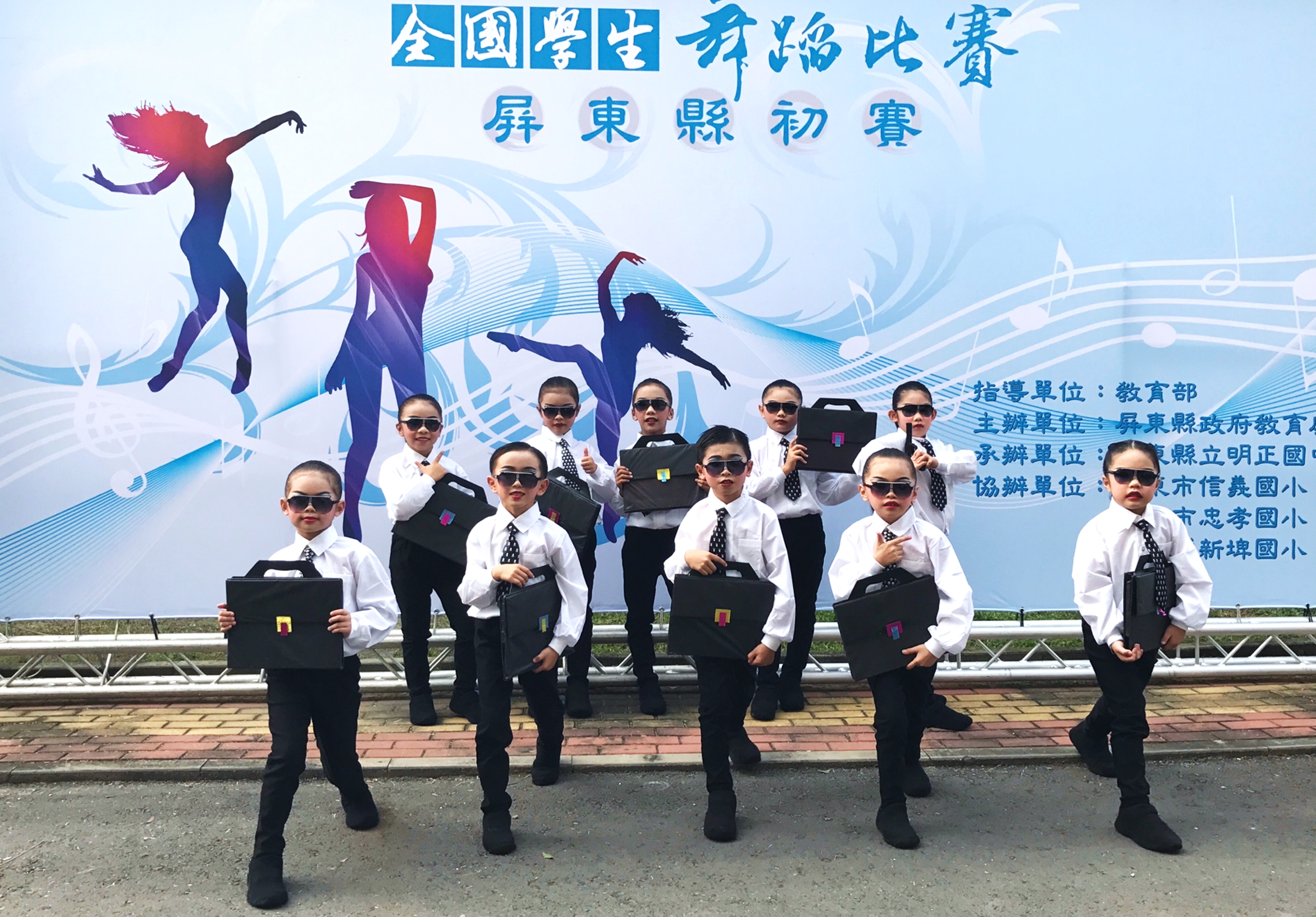 全國學生舞蹈比賽屏東縣初賽海報前，約十名身穿正裝、戴墨鏡的學生排成一列，手持黑色公事包擺出舞蹈姿態。背景為藍色漸層設計，搭配音樂符號和舞者剪影圖案。