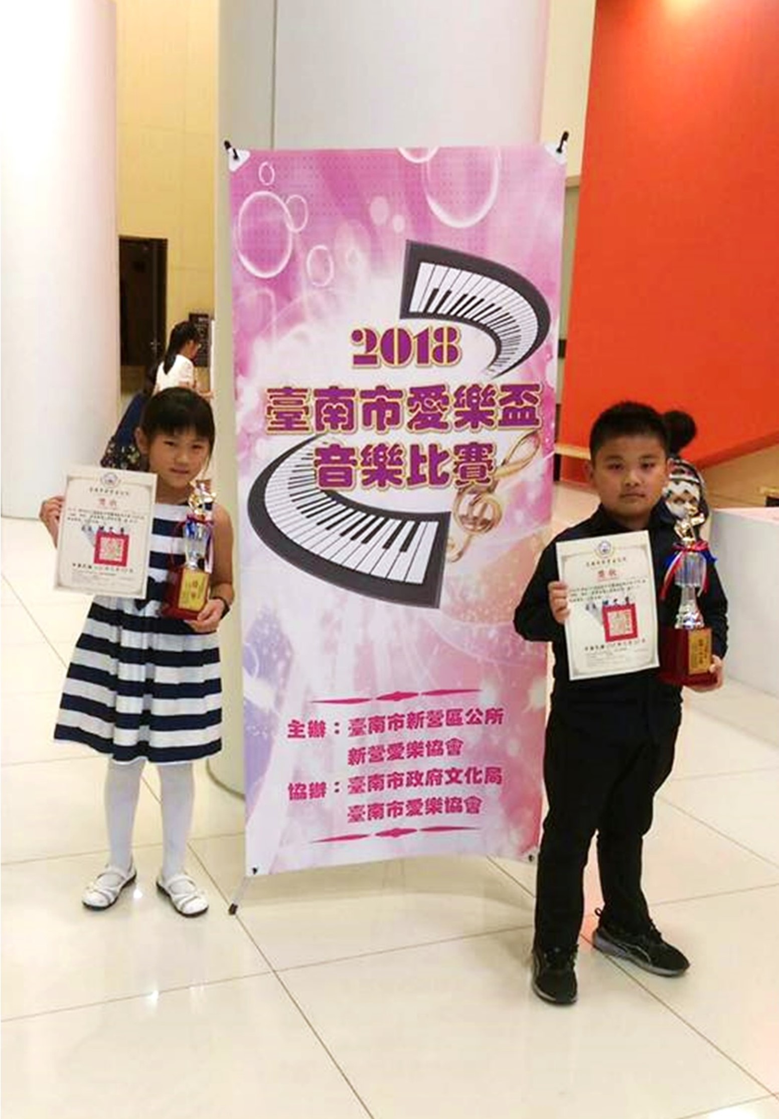 兩名小女孩在2018臺南市愛樂盃音樂比賽會場前合影，各自手持獲獎證書和獎杯。背景為粉紅色海報，上有鋼琴鍵盤圖案和賽事相關資訊。