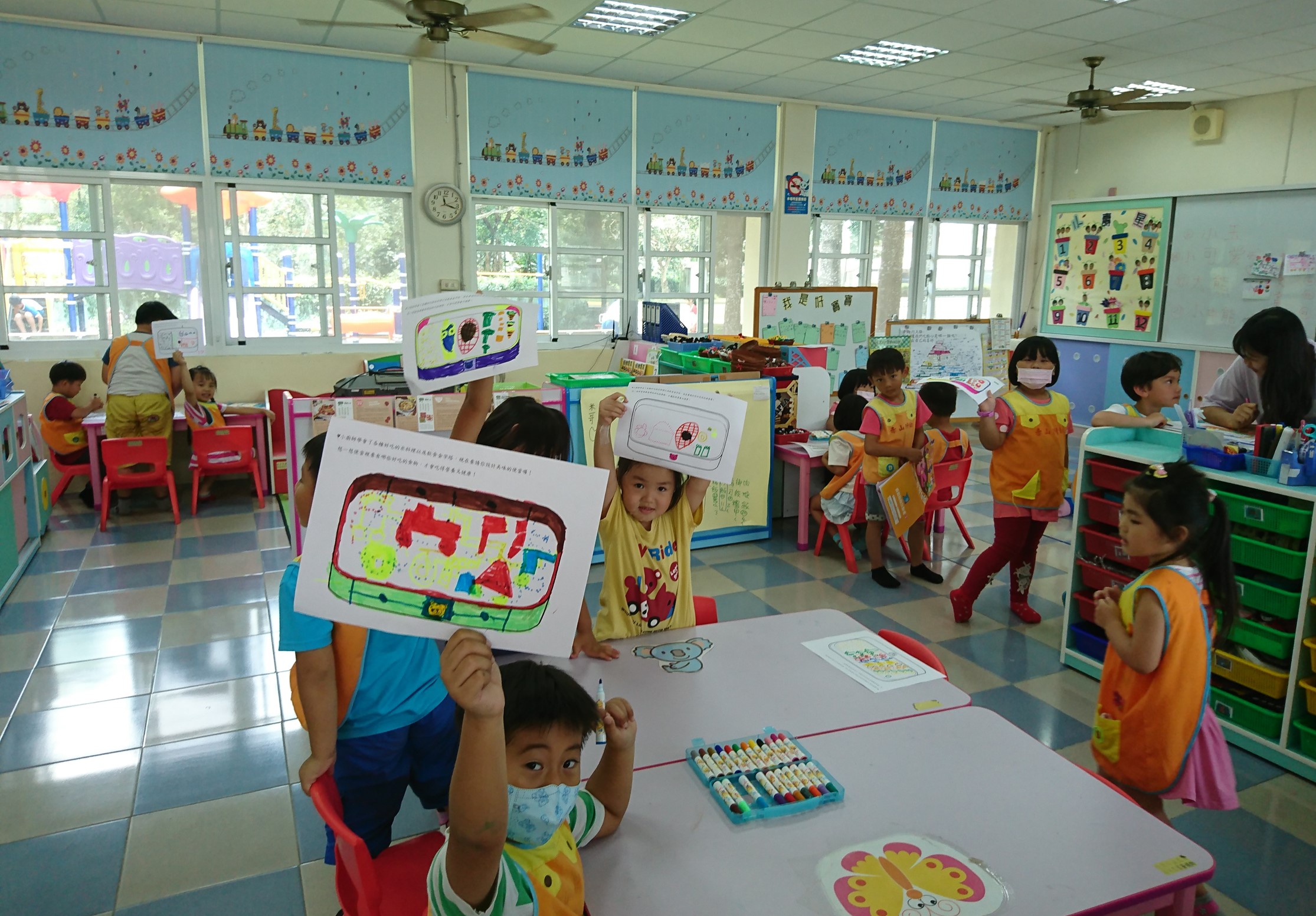 幼兒園教室內，多名小朋友穿著橙黃色背心展示自己的繪畫作品。教室牆面貼滿彩色裝飾，窗邊放置紅色小椅子和桌子，地板為彩色方格磁磚。孩子們在進行美術創作活動，展示各自完成的畫作。