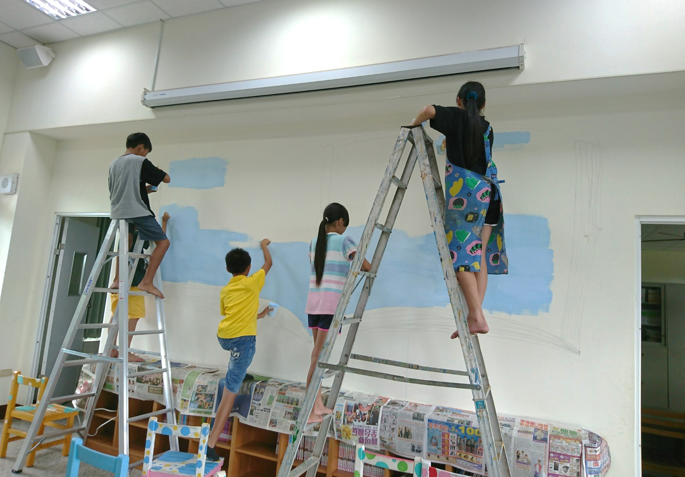 四名兒童在教室裡站在梯子上，共同繪製牆上的彩畫。牆面鋪著報紙保護，已畫出藍色天空和米色地面的背景。兒童穿著不同顏色衣服，正專注地進行牆面彩繪創作活動。