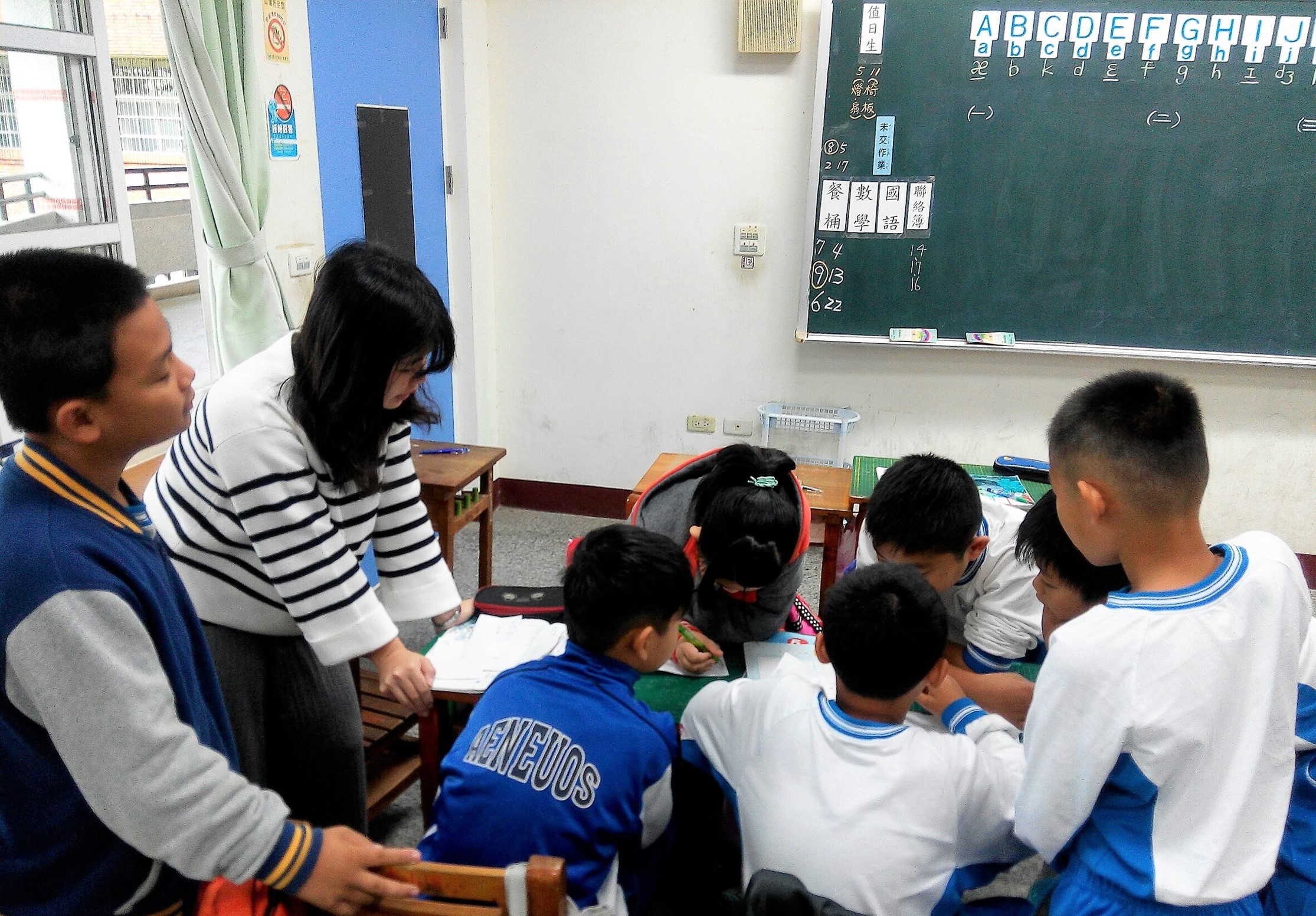 教室內，一位女老師和一位男性助教正在指導約六名小學生進行英文學習。學生們圍坐在一起，黑板上顯示英文字母和中文字，教室窗邊可見綠色窗簾。