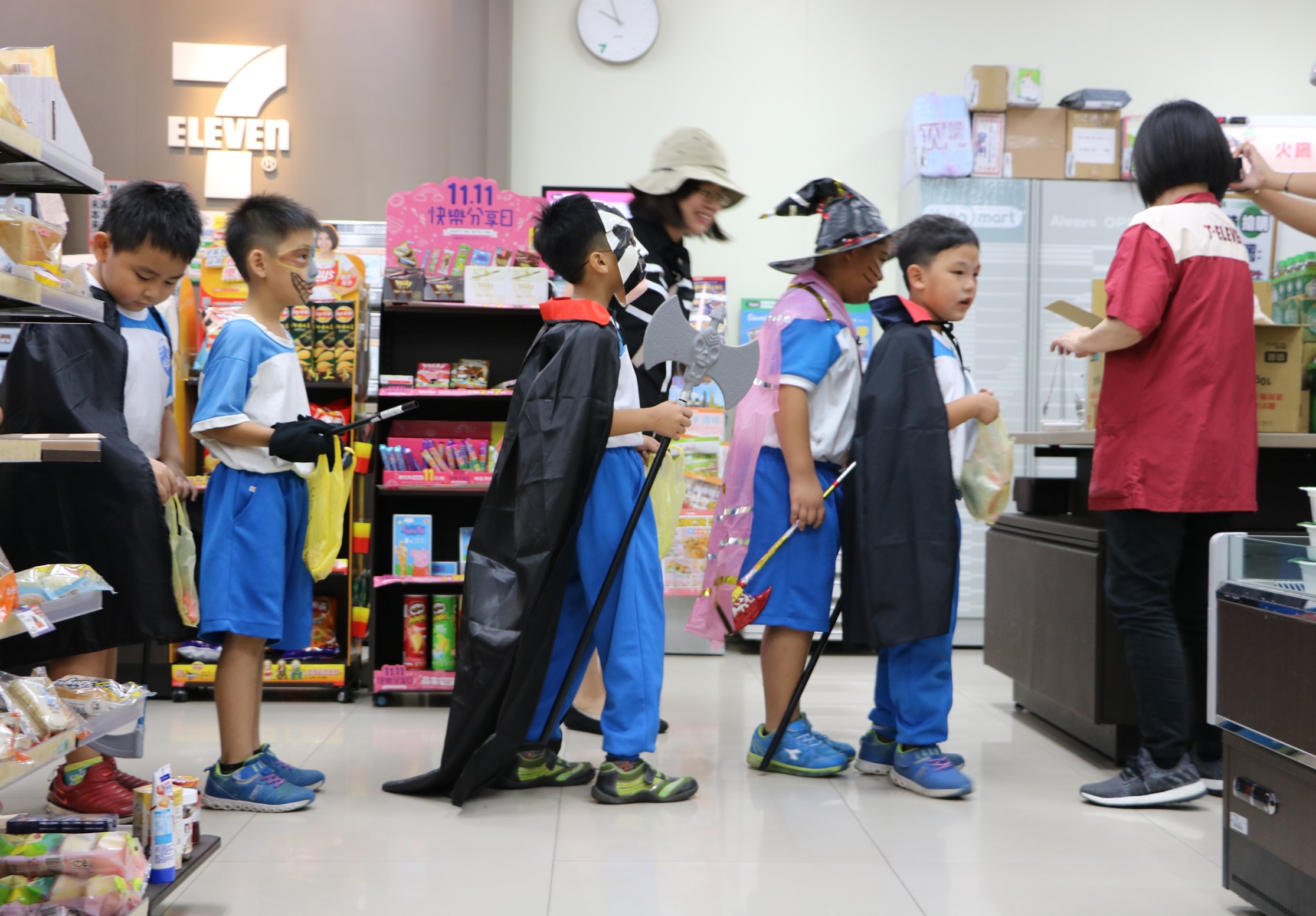 一群小學生在便利商店內進行萬聖節變裝活動，穿著各式童趣服裝，包括巫師、吸血鬼等角色扮演，手持道具，在店內走動。背景可見7-ELEVEN標誌和商品陳列架。