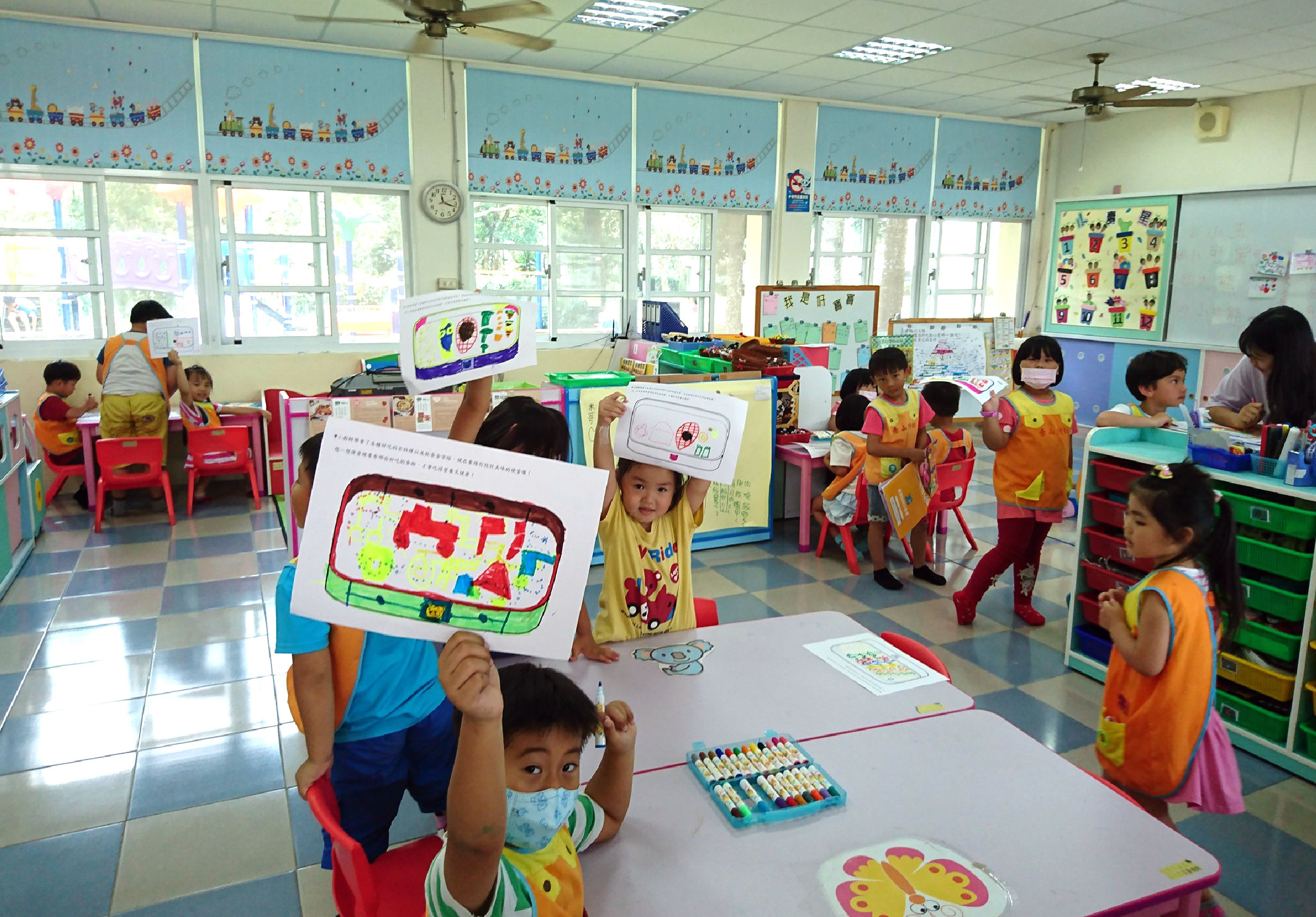 幼兒園教室內，多位穿著黃色背心的小朋友展示自己的繪畫作品。教室設有五彩繽紛的傢具、美術用品和教學玩具。窗戶上裝飾著列車和花卉圖案的布置。小朋友們在進行美術創作活動。