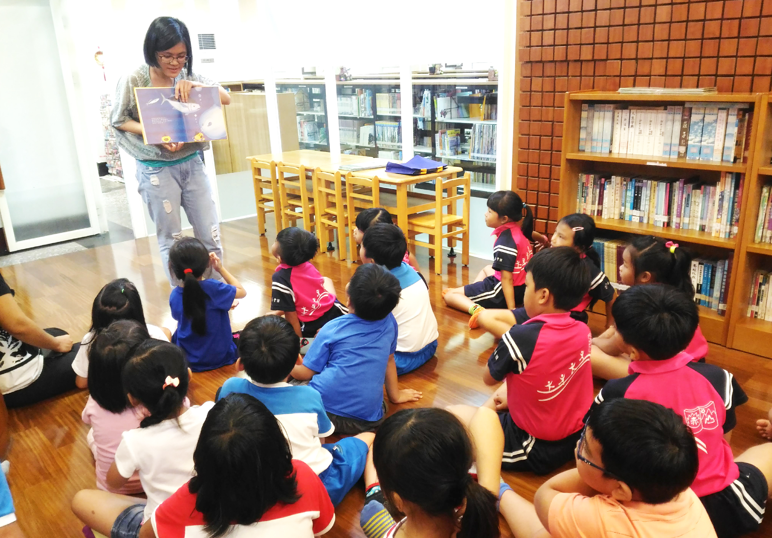圖書館內，一位女性講師向坐成圓圈的二十多名小學生展示繪本，進行說故事活動。孩子們穿著紅藍色運動服，認真聆聽。背景可見書架、桌椅和借閱櫃台。
