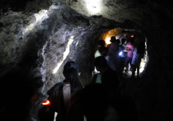 一群遊客在地下洞穴內參觀，手持手電筒和照明設備照亮岩石壁面。洞穴內光線昏暗，岩石紋理清晰可見，人們聚集在狹窄的通道中，進行洞窟探險活動。