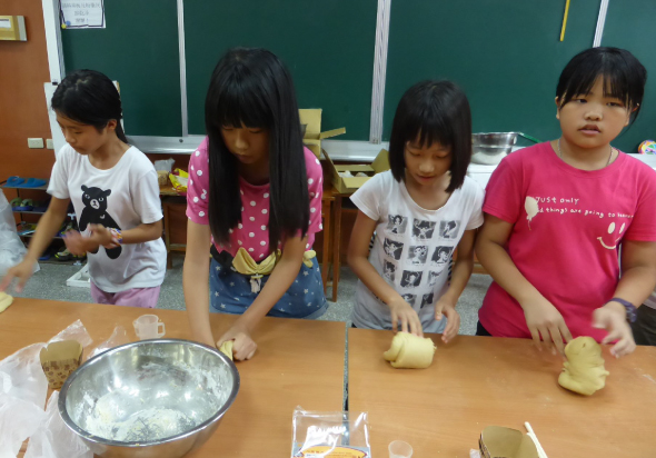 四位小女孩在教室裡進行烹飪課程。她們站在桌前準備食材，面前有一個大碗、馬鈴薯和其他烹飪工具。背景是綠色黑板和教室環境。孩子們穿著彩色T恤，認真地參與這項手作食物活動。