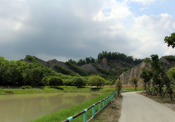 河邊風景，綠色鐵欄杆護欄沿著平坦的白色步道延伸，河水呈褐色，兩岸被茂密的綠樹環繞，遠處可見陡峭的岩壁和山丘，天空有白雲點綴。