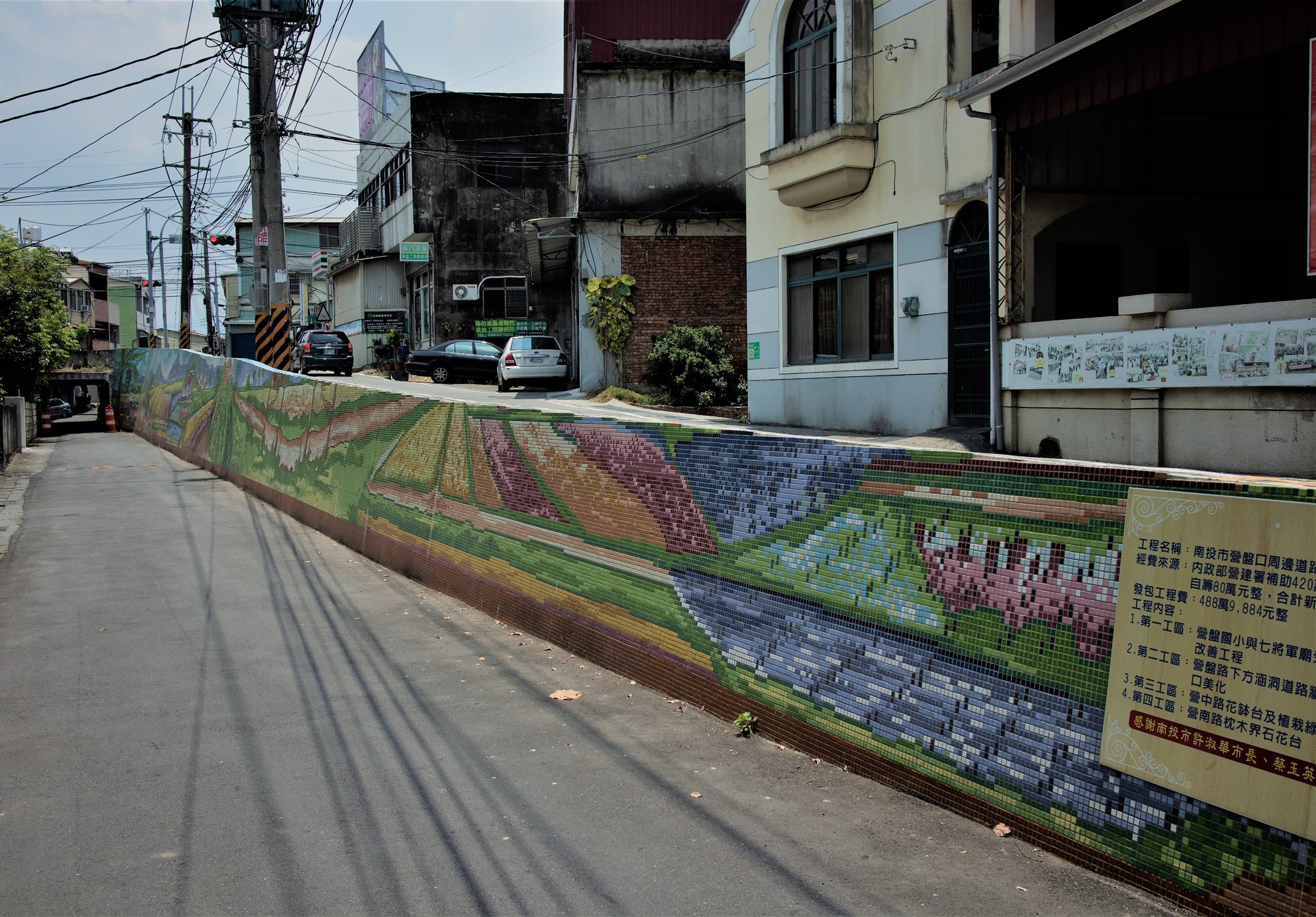 台灣街道巷弄內，一面彩色馬賽克磁磚牆面描繪農田風景，有綠色稻田、紫色花卉等圖案。周圍是老舊建築、停放車輛、電線桿等都市景觀。牆面右側有工程說明牌。