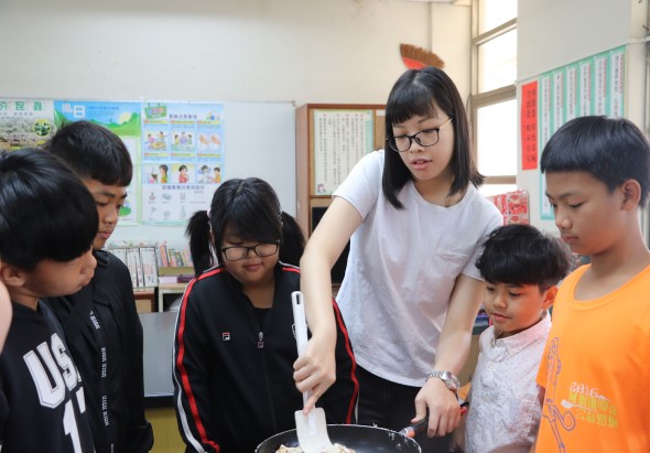 教室內，一位女性教師穿著白色T恤，正在指導圍成一圈的六位學生進行科學實驗。學生們穿著不同顏色的衣服，專注地觀看老師演示操作。教室背景有教學海報和書籍。