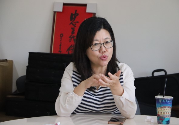 一位戴眼鏡的女性坐在辦公室內，穿著白色外套和黑白條紋內衣，雙手做出說話手勢。身後牆上掛著紅色中文書法作品，桌上放著飲料杯，似乎在進行簡報或講座。