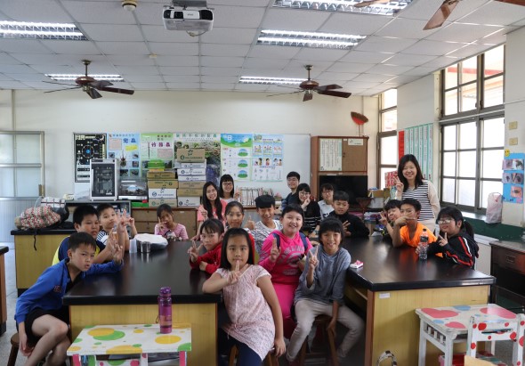 小學教室內，一位女教師站在講台旁，約三十名小學生圍坐在黑色課桌周圍，認真聆聽上課。教室牆面貼有教學海報和學生作品，天花板裝有風扇和投影機，窗戶採光充足。