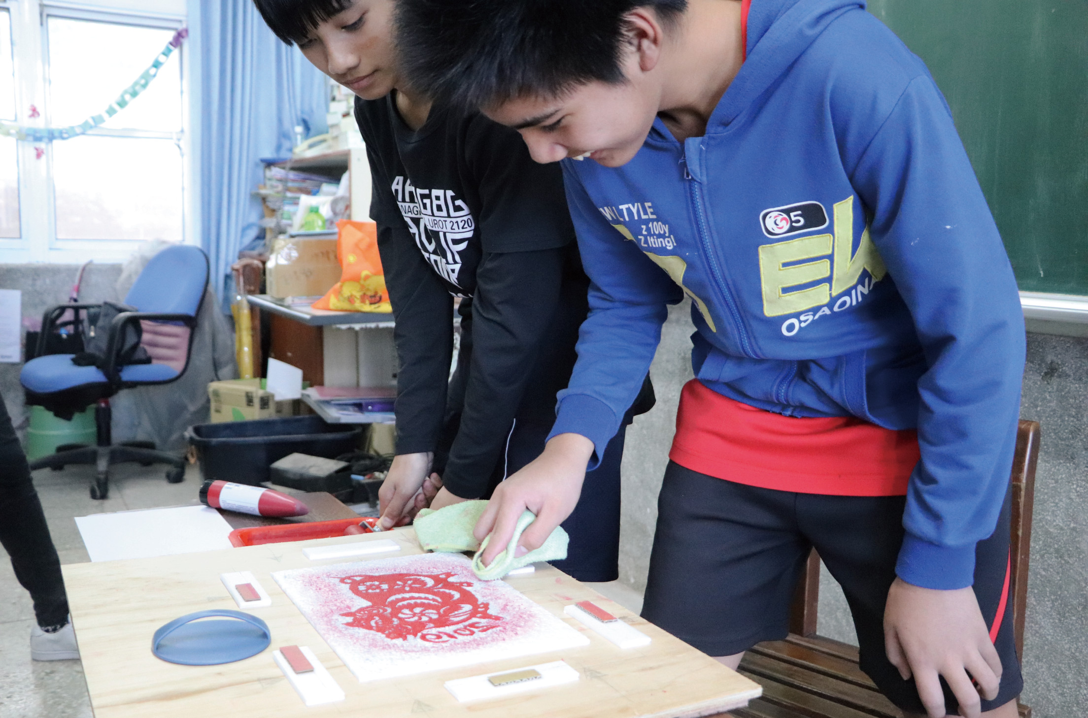 三名學生在教室內進行美術活動，在木製工作台上製作紅色剪紙藝術作品。左側學生穿著黑色上衣，中間和右側學生穿著藍色運動服。他們專注地用綠色工具在紙上進行創作，桌上擺放著紅色蠟筆和其他美術用具。