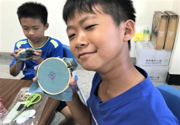 兩名穿著藍色制服的男童在教室內進行刺繡手工藝活動。前景的男童展示完成的藍色圓形刺繡作品，後景的男童正在進行刺繡。桌上擺放剪刀、線材和其他手工用具。背景可見書籍和教室物品。