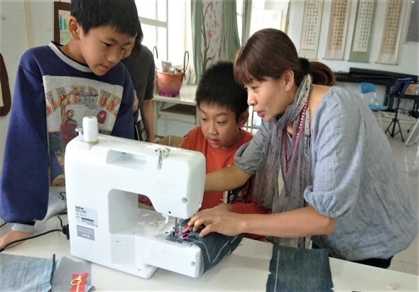 一位女性教師在教室裡教導三名小學生使用縫紉機。學生穿著校服，認真觀看並學習操作機器。教室背景擺放著教學用具和掛圖。這是一堂手工藝技能課程。