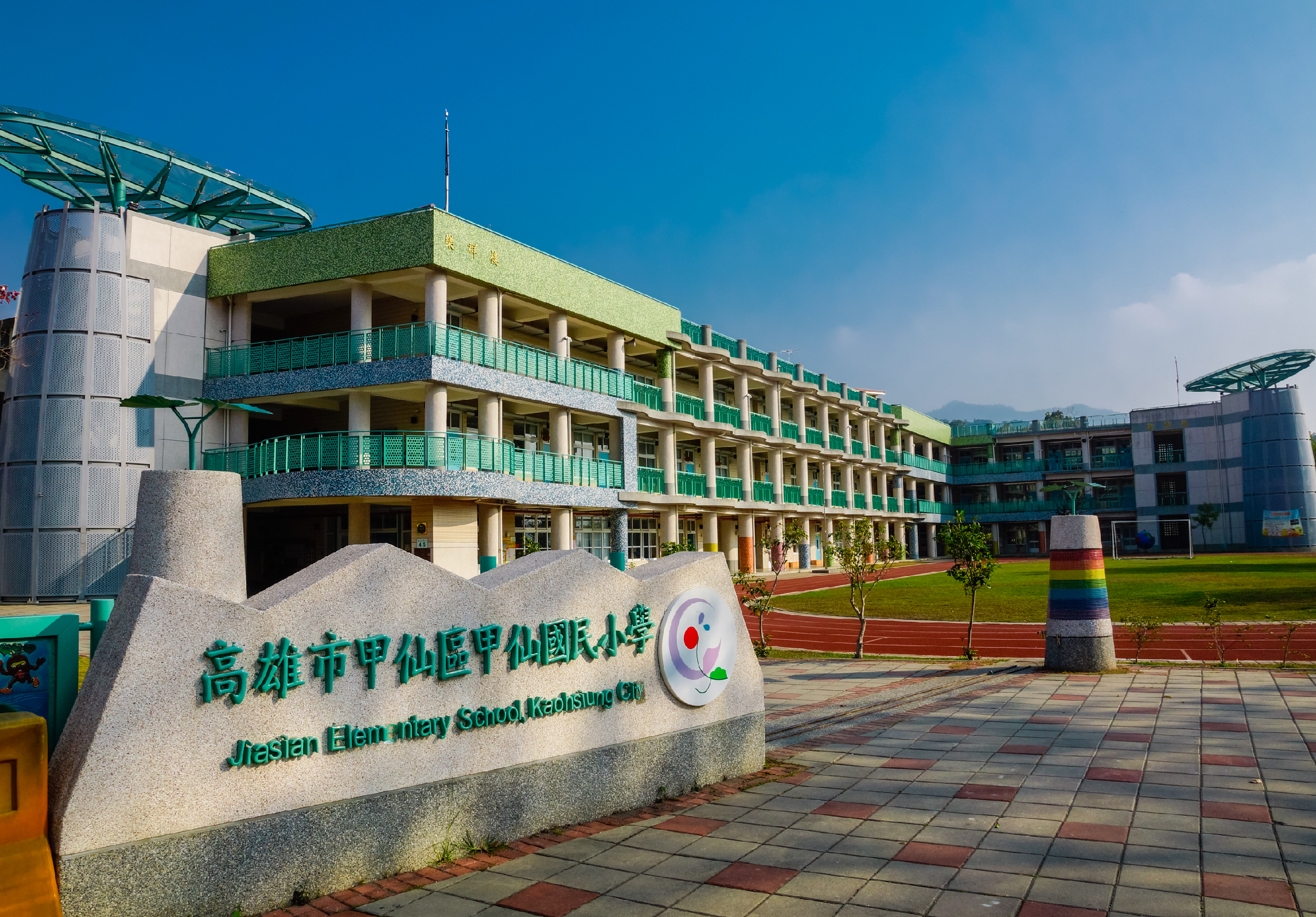 高雄市甲仙國小校園全景，三層樓綠色欄杆教學大樓，紅色跑道運動場，校名石碑立於前方，晴朗藍天，遠方可見山景。
