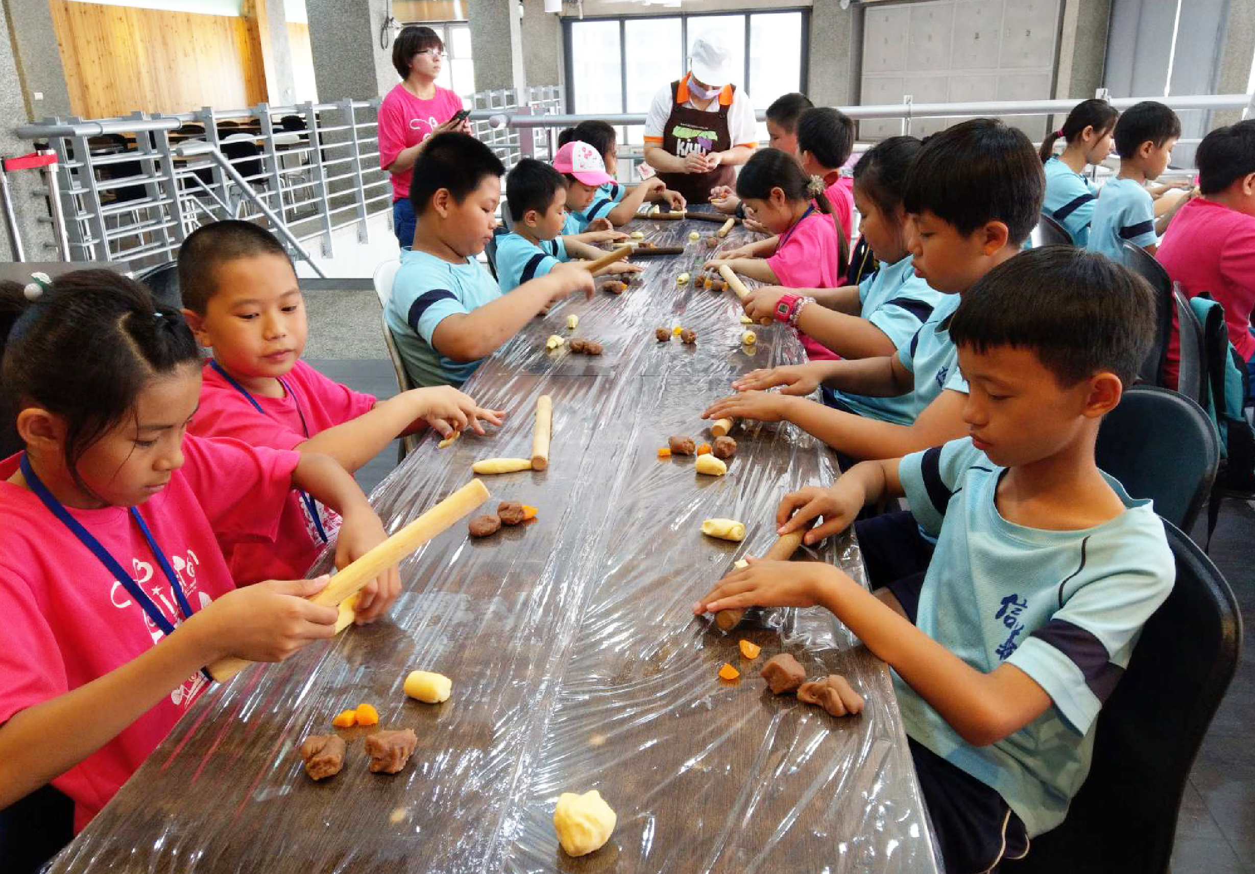 一群穿著粉紅色和藍色衣服的小學生，在室內長桌旁進行食物加工活動。他們用手製作或處理桌上的食材，包括玉米粒和麵包等。背景有成人指導，場景是校園或教室環境的集體學習活動。
