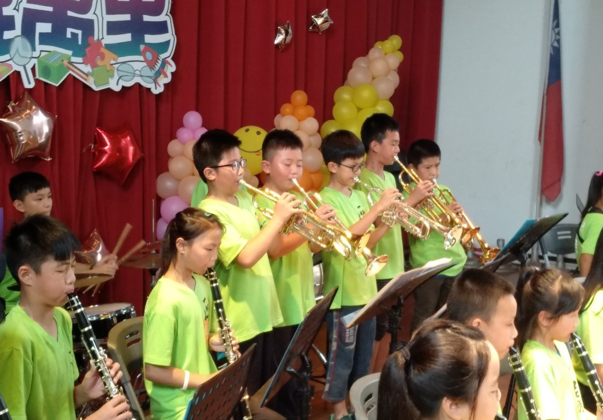 一群穿著螢光綠色制服的兒童在舞台上進行樂器表演。背景是紅色窗簾，裝飾著彩色氣球和金色星星。孩子們正在演奏小號、豎笛等樂器，進行音樂會表演。