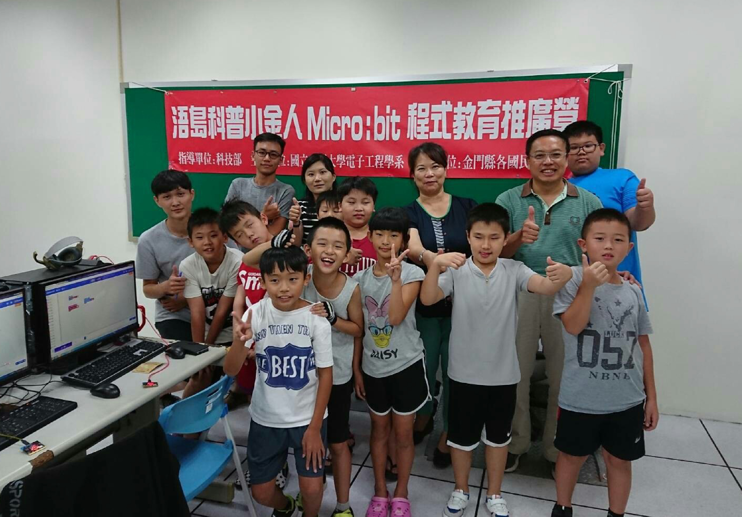 一群學生和教師在教室內合照，背後是紅綠相間的海報，上面標示Micro:bit程式教育推廣字樣。前排學生做出勝利手勢，後排成人站立，左側可見電腦和鍵盤，氣氛歡樂友善。