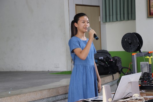 穿著淺藍色連衣裙的女性在室內舞台上拿著麥克風進行演講或主持。身旁放有筆記型電腦和其他設備。背景為白色牆壁、綠色線簾和風扇，是一場講座或活動現場。